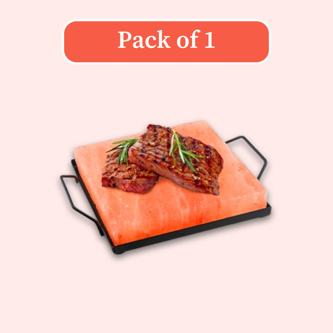 A rectangular pink salt stone serving tray with black handles, shown with two grilled steak slices on top and rosemary garnish, under a coral 'Pack of 1' label on a pale pink background.