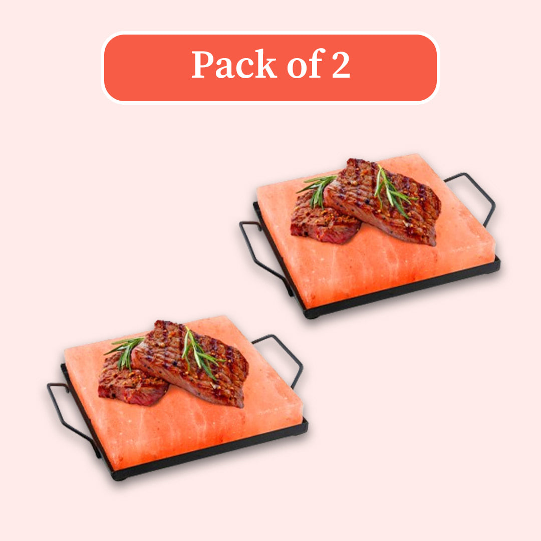 Two rectangular pink salt stone plates with black metal handles, each topped with grilled steak and a sprig of rosemary, shown on a pale pink background with an orange label reading 'Pack of 2'.